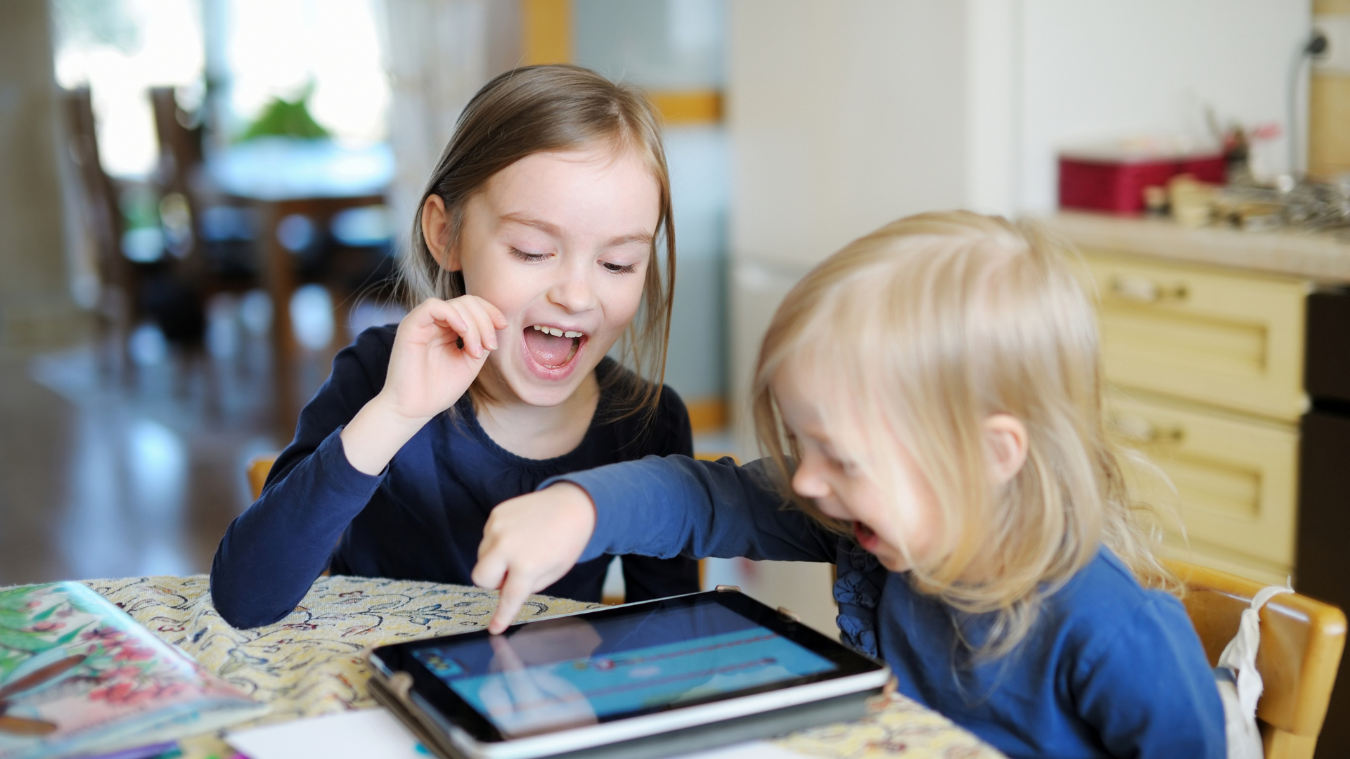 Two young girls playing together on a tablet at a table. The younger child is pointing at the tablet and both have their mouths open in laughter.