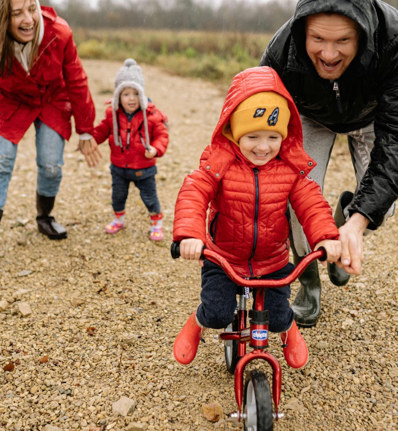 Dad helps young son learn to ride bike while mum and younger brother watch in the background.