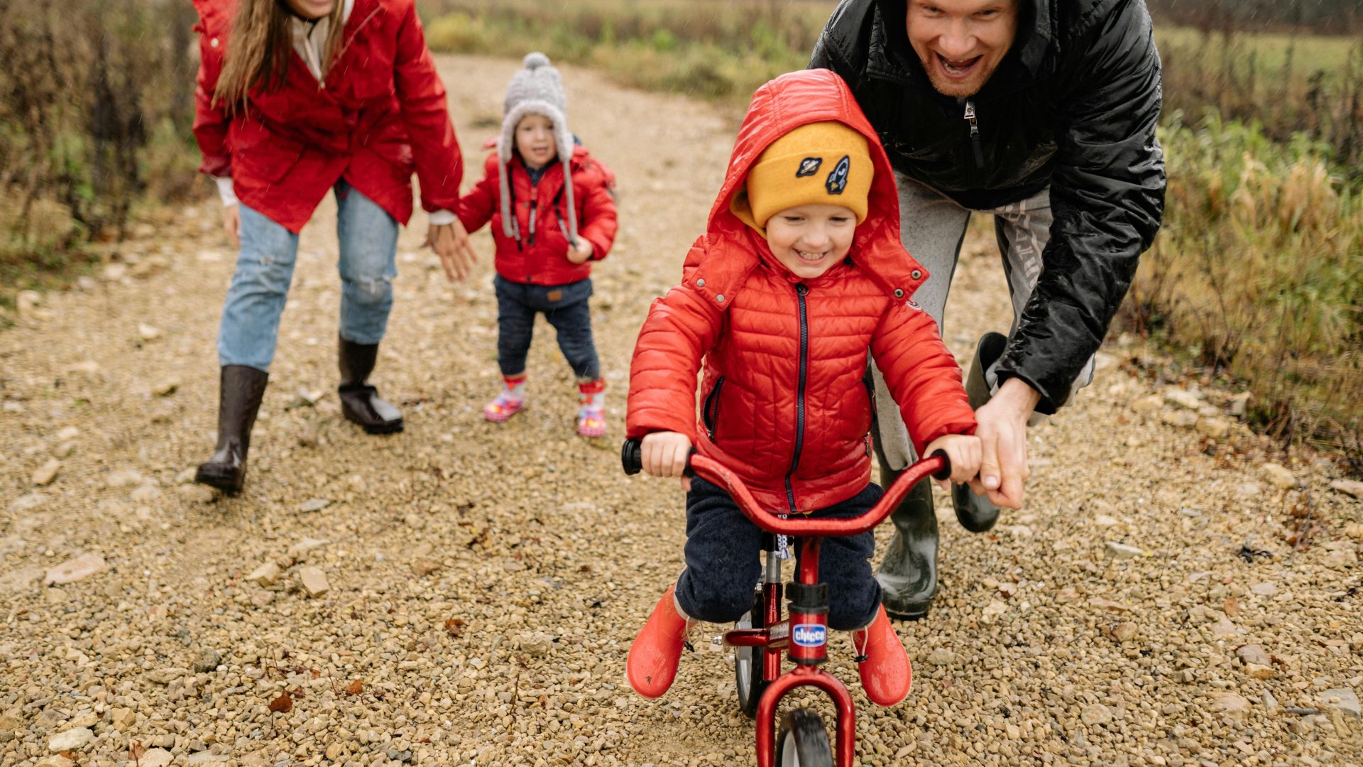 Dad helps young son learn to ride bike while mum and younger brother watch in the background.